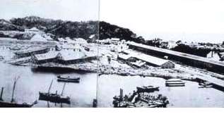 Photographie ancienne de l'arsenal de Yokosuka : baie, digue, hangars et embarcations au bord de l'eau