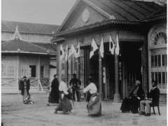 Démo (Ono Ha Itto Ryu) Photographie ancienne d'un bâtiment japonais de style occidental pavoisé de bannières, passants en kimono devant